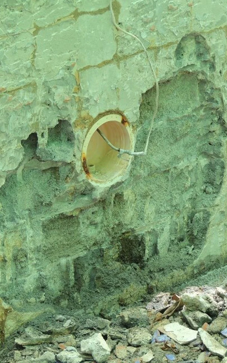 A round wall fitting set into an empty pool wall, with a cable threaded out through its opening, rust staining down the inside of the ring and brown staining on the wall around it, the surrounding surface layer flaked and lifted away to the bare gritty layer beneath, stained horizontal cracks running across the wall above, and broken render and tile fragments banked against the base.