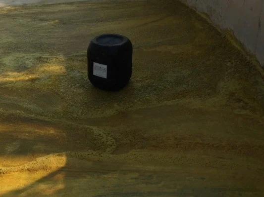 A black drum with a white label stands on a wet floor stained green and yellow, beside a grey wall. Sunlight falls across the lower left of the floor.