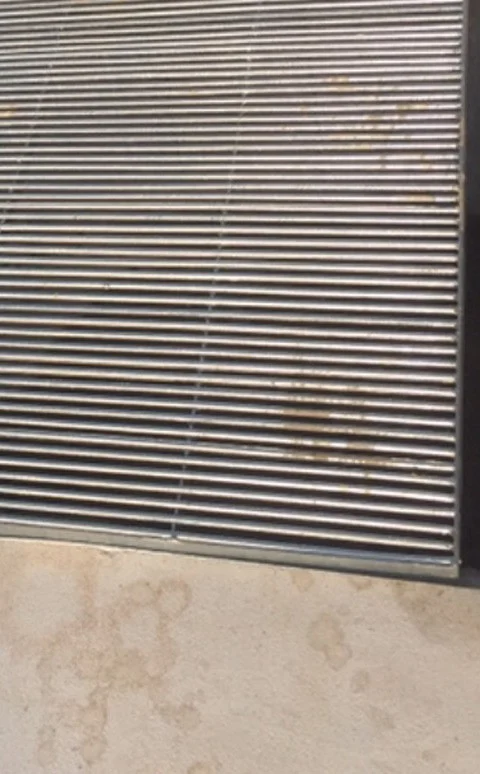 A linear metal grating of closely spaced pale bars, several of them carrying ochre-brown deposits in specks and short streaks, with a plain pale deck below it marked by brown blotching, overlapping circular ring marks and dried drip trails.