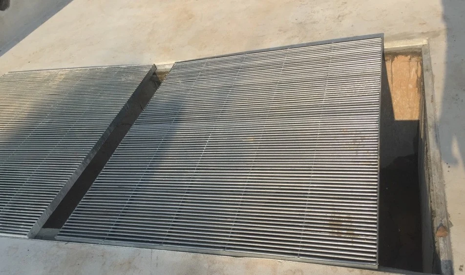 Steel bar-grating panels laid over a rectangular opening in a pale floor. The right-hand panel stops short of the rim on that side, leaving part of the opening uncovered, and a tapered slot runs between the two panels, so the dark void below shows through both gaps. Brown staining runs down the grating beside the uncovered section, and the floor and the rim around the opening carry dark smears, brown discolouration and loose dirt.