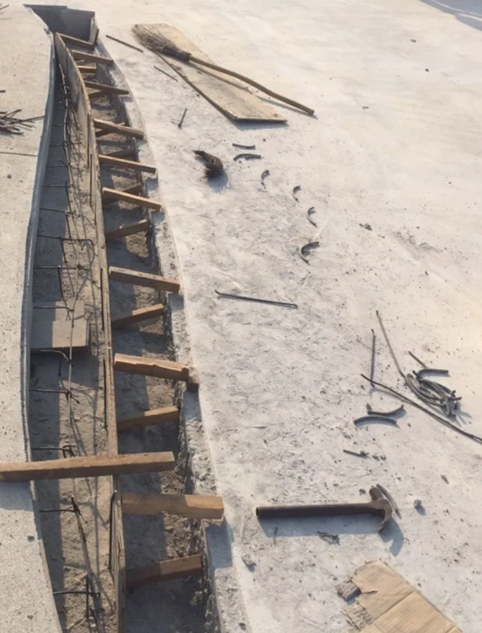 A flat, dusty grey surface scattered with bent steel offcuts, loose steel rods, a claw hammer, a broom lying on a loose timber board and a sheet of cardboard, beside a long curved channel holding steel reinforcing bars and an upright timber board propped with short timber struts.