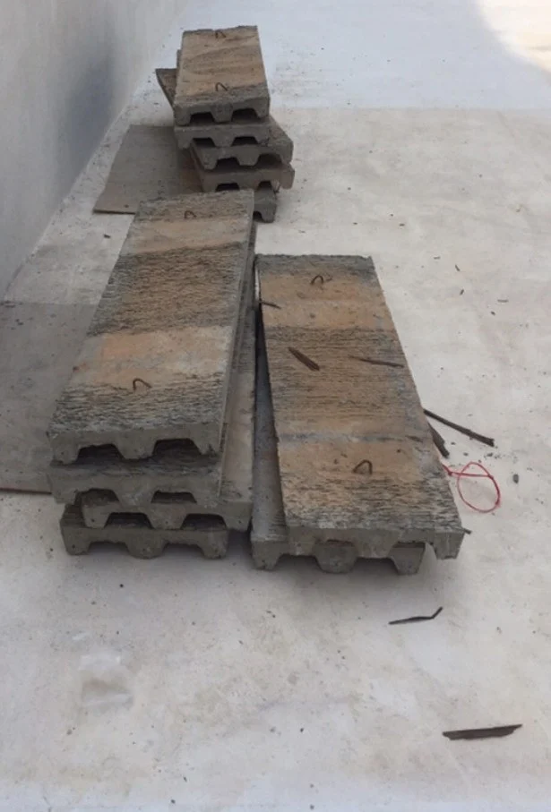 Concrete planks with ribbed undersides and small wire loops on top sit stacked on a smooth pale grey floor beside a tall wall, with thin boards under only part of the load, the nearest planks resting directly on the floor, and dark scraps and a red loop lying loose beside them.