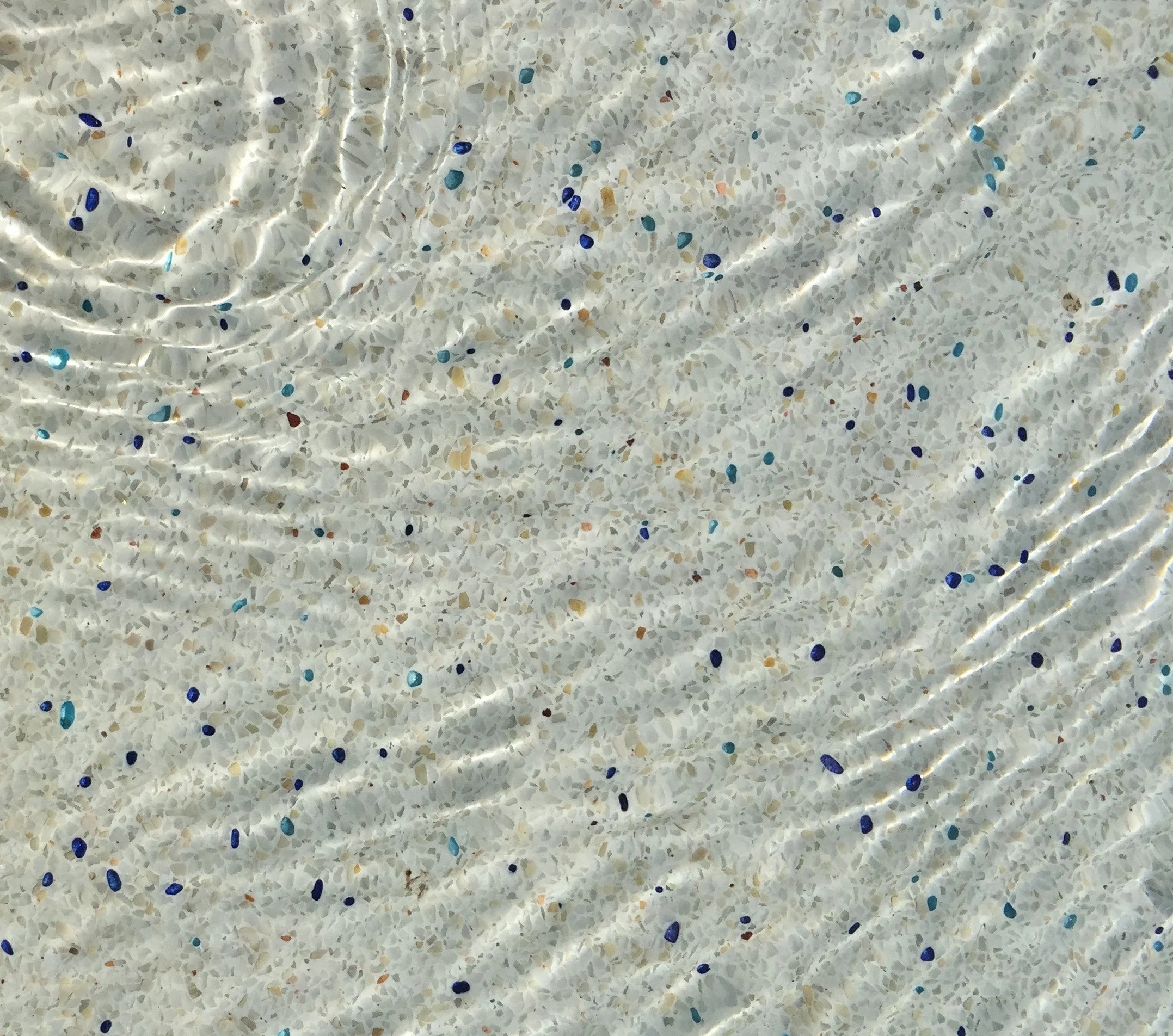 A pool floor in white exposed aggregate, seen from above through clear water, with pale stone plus amber, rust, blue and turquoise glass crystals standing proud of a white matrix and ripple light crossing the surface.