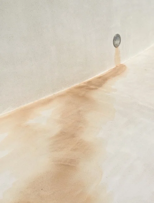 A rust-coloured streak runs from a small round light fitting set in a pale pool wall down to the junction with the floor, where a broad tan stain has dried in a fan across the dry pale surface.