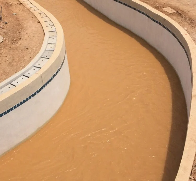 A curved pool channel stands part-full of opaque orange-brown water, with smooth pale walls, a dark tile band below the coping, and bare loose earth and scattered rubble running up to both edges.