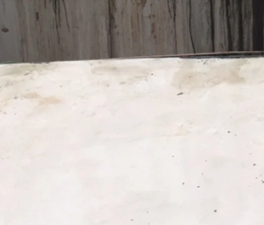 Close view of the face of a pool wall meeting the pale floor beside it. The wall runs off the top of the frame and is streaked the whole way down with dried grey runs and a few small rust-brown blotches, the streaking deepening almost to black towards the right-hand end. A dark shadow line marks where the wall meets the floor, with a narrow lit edge above it along the right-hand half. Below, the pale, chalky floor slab is smudged grey-brown along the base of the wall and scattered with small dark flecks further out.