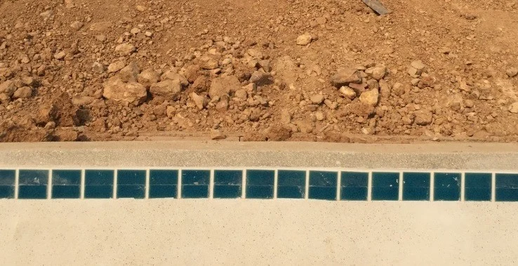 A heap of loose reddish earth and broken stone banked up against the pale surround of an empty pool, with soil and dust lying on the surround, a granular exposed-aggregate edge band below it, and a single row of dark teal waterline tiles below that, dry with no water against them.