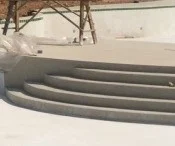 Curved steps lead down into an empty pool. Timber props stand on the higher ground behind them, in front of a white wall.
