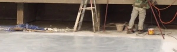 A smooth pale grey pool floor. Along its back edge are a blue tarp, a line of rubble, a stepladder, red hoses, a bucket and a yellow paint tin, and a worker's legs and boots stand on the floor.