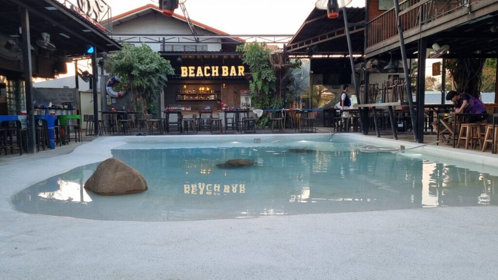 Wide daylight view across a freeform pool towards the Beach Bar counter and surrounding seating.