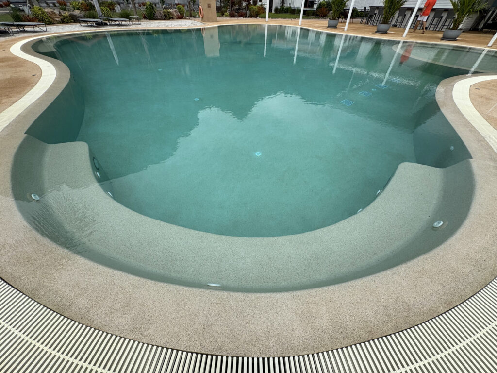 Elevated view across the freeform pool at This Great Place Resort, showing curved submerged seating and the continuous aggregate finish.