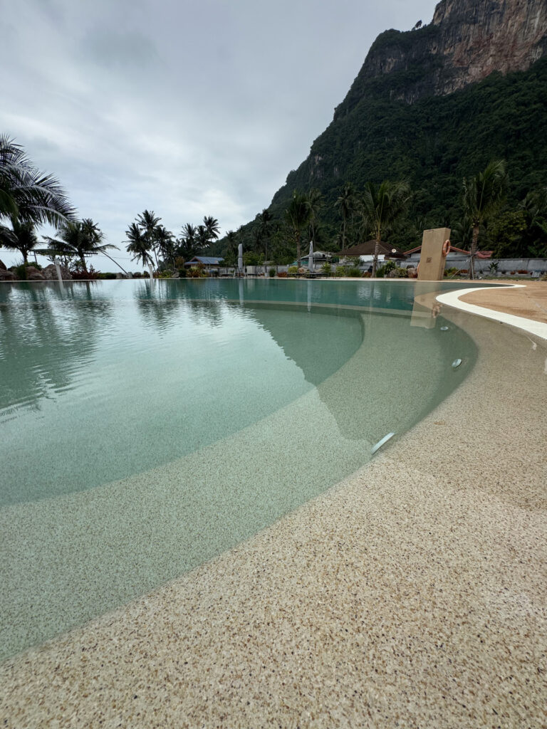 View along the curved pool edge and submerged seating at This Great Place Resort, with palm and mountain reflections on the water.