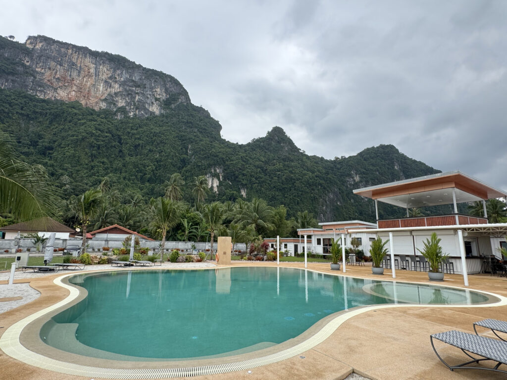 Wide view of the curved pool at This Great Place Resort with landscaped grounds, a poolside building and a forested mountain backdrop.