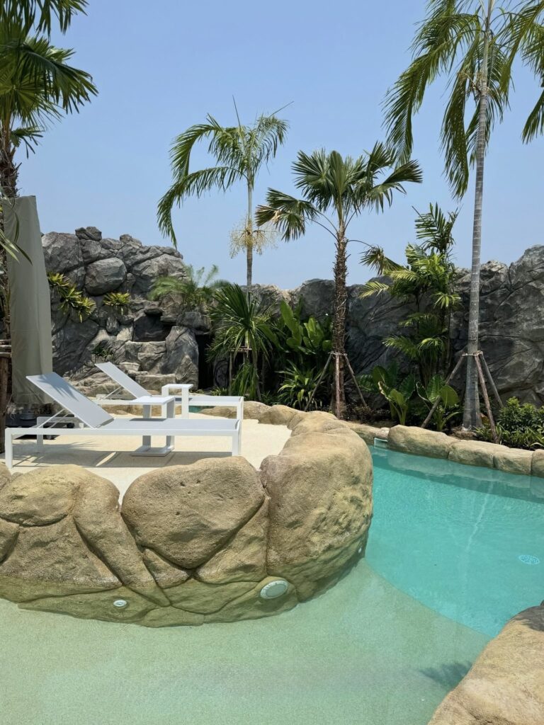 Pale pool finish beneath shallow water beside a rock-edged island with white sun loungers.