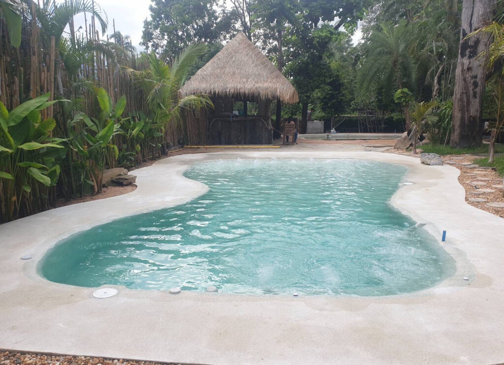 Wide view of a freeform pool surrounded by a pale finish, palms, bamboo and a thatched garden pavilion.