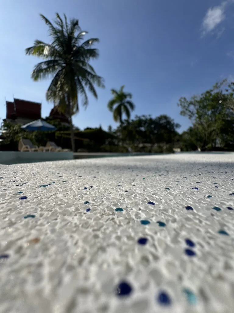 Close-up of pale aggregate with blue and turquoise flecks, with the pool and palms softly blurred behind.