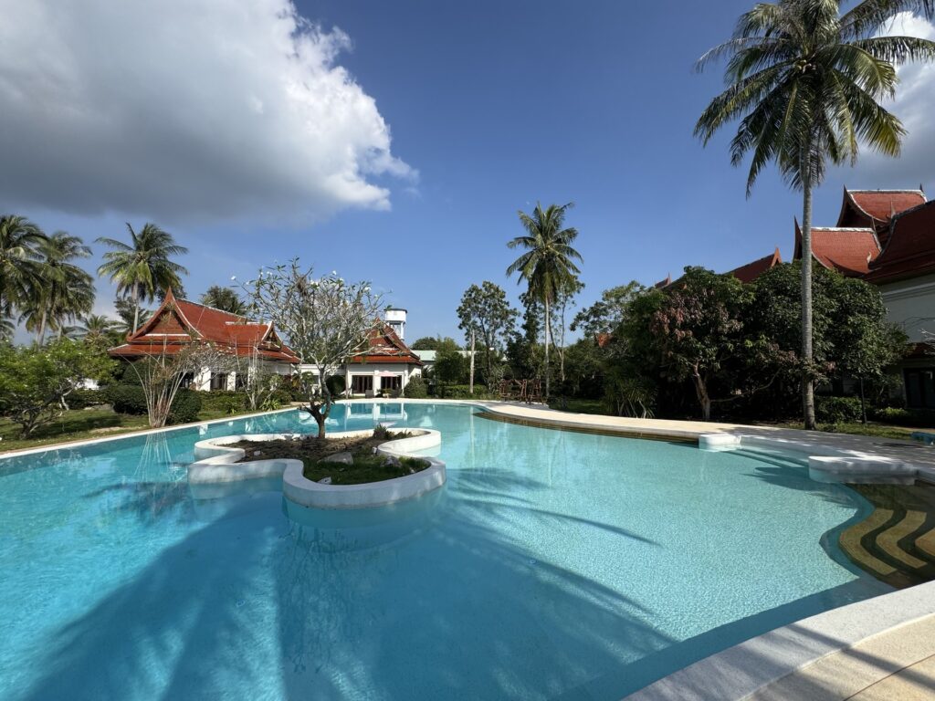 Wide view of a shared lagoon pool with a planted island, palms and Thai-style villas in Phuket.