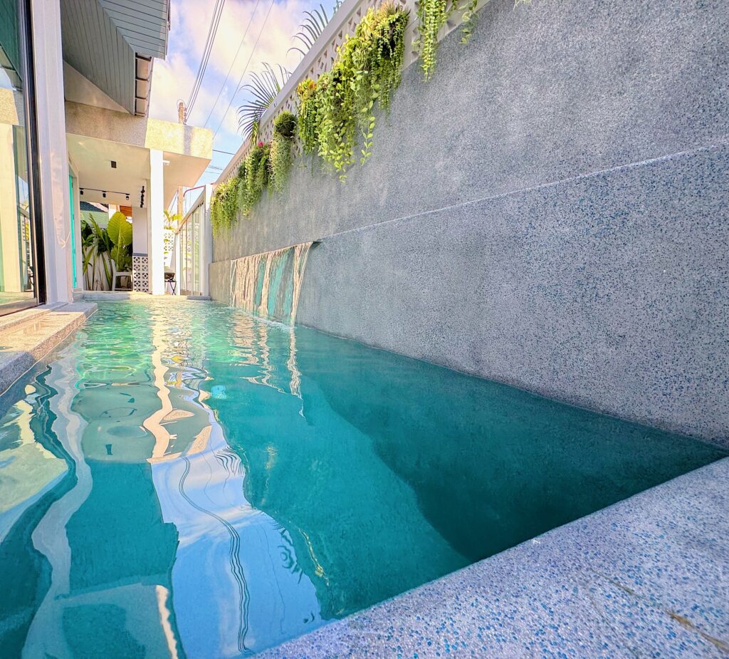 A narrow lap pool seen along its length, with exposed-aggregate coping in the foreground, a grey exposed-aggregate wall on the right with water spilling from it in a sheet, and the house reflected in the rippled surface.