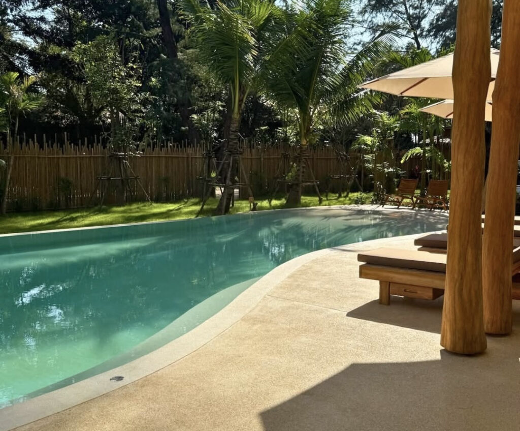 Poolside view at The Talay showing the curved Pearl White pool edge beside shaded loungers and a tropical garden.